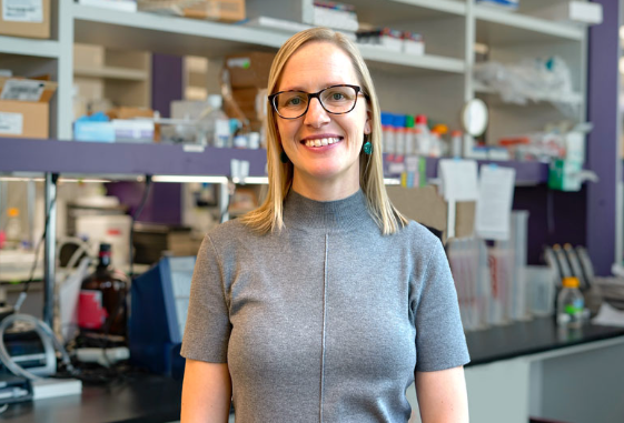 A woman wearing glasses and a grey shirt smiles in a lab